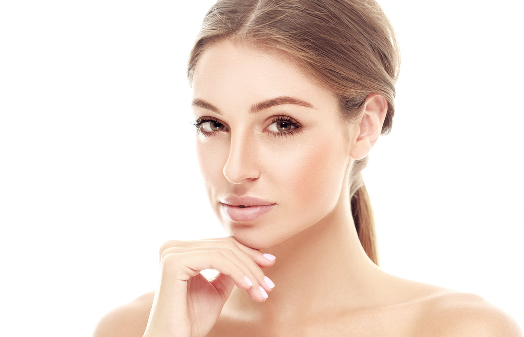 A woman with light brown hair is looking at the camera with her hand touching her chin. The background is white.