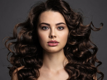 Woman with long, curly brown hair and neutral expression against a gray background.