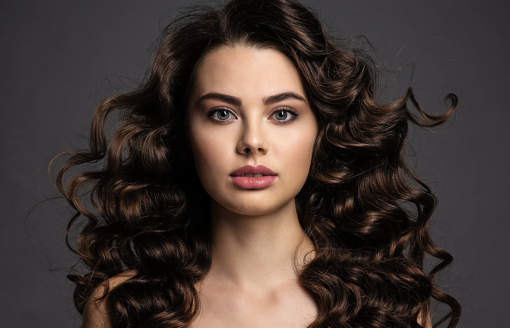 Woman with long, curly brown hair and neutral expression against a gray background.