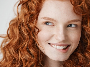 Person with wavy red hair smiling and looking to the side against a plain background.