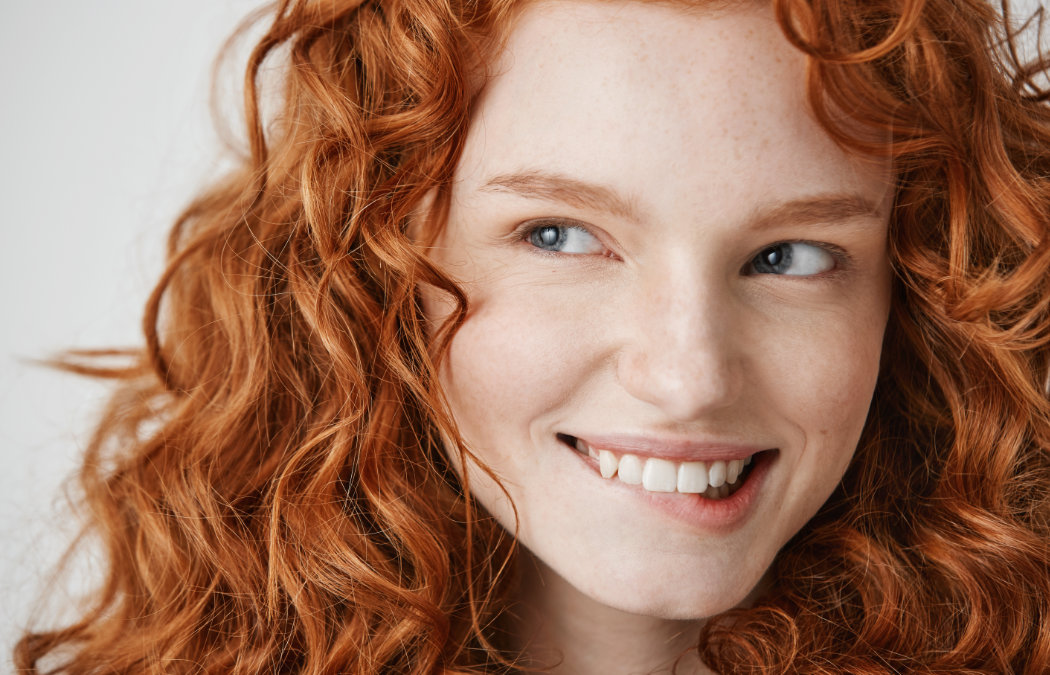 Person with wavy red hair smiling and looking to the side against a plain background.